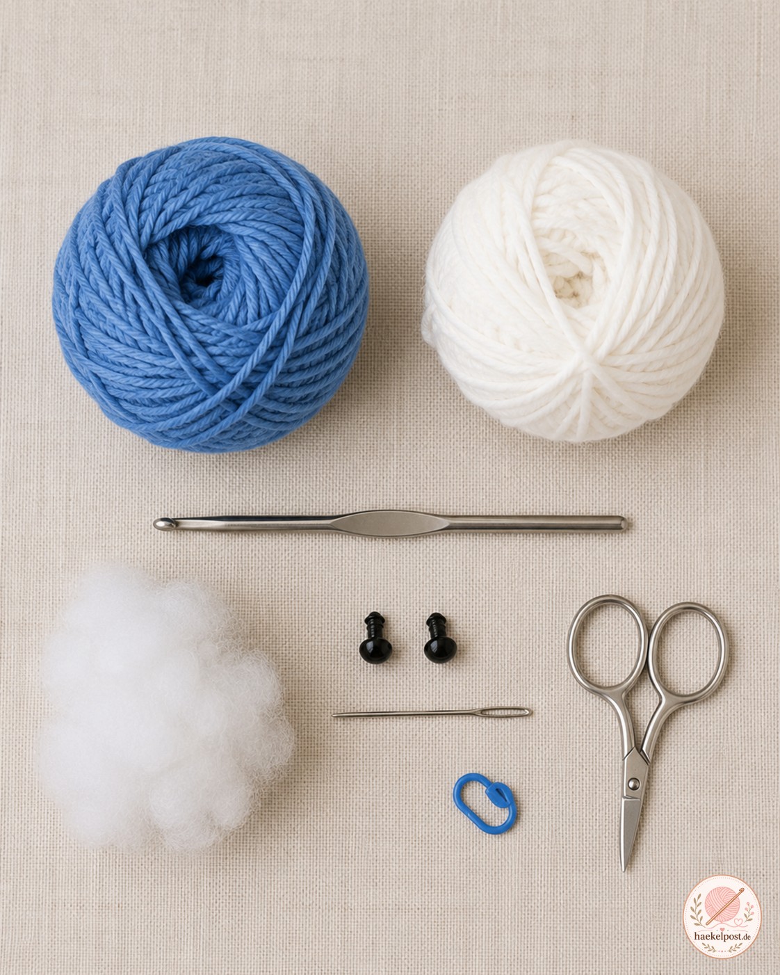 Material-Flatlay zum Wal häkeln mit blauem und weißem Garn, Häkelnadel und Sicherheitsaugen auf Leinen.